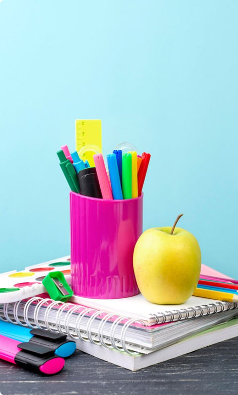 A desk with stacked notebooks, a pink pen holder filled with colorful pens, and a green apple