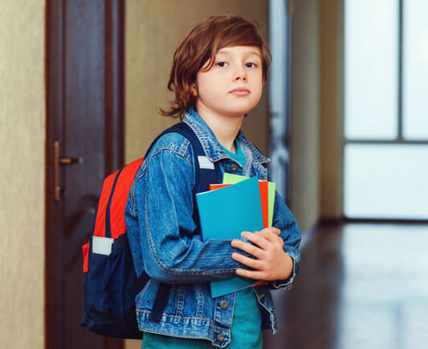 A student with a serious expression holding books and wearing a denim jacket and backpack, standing in a hallway